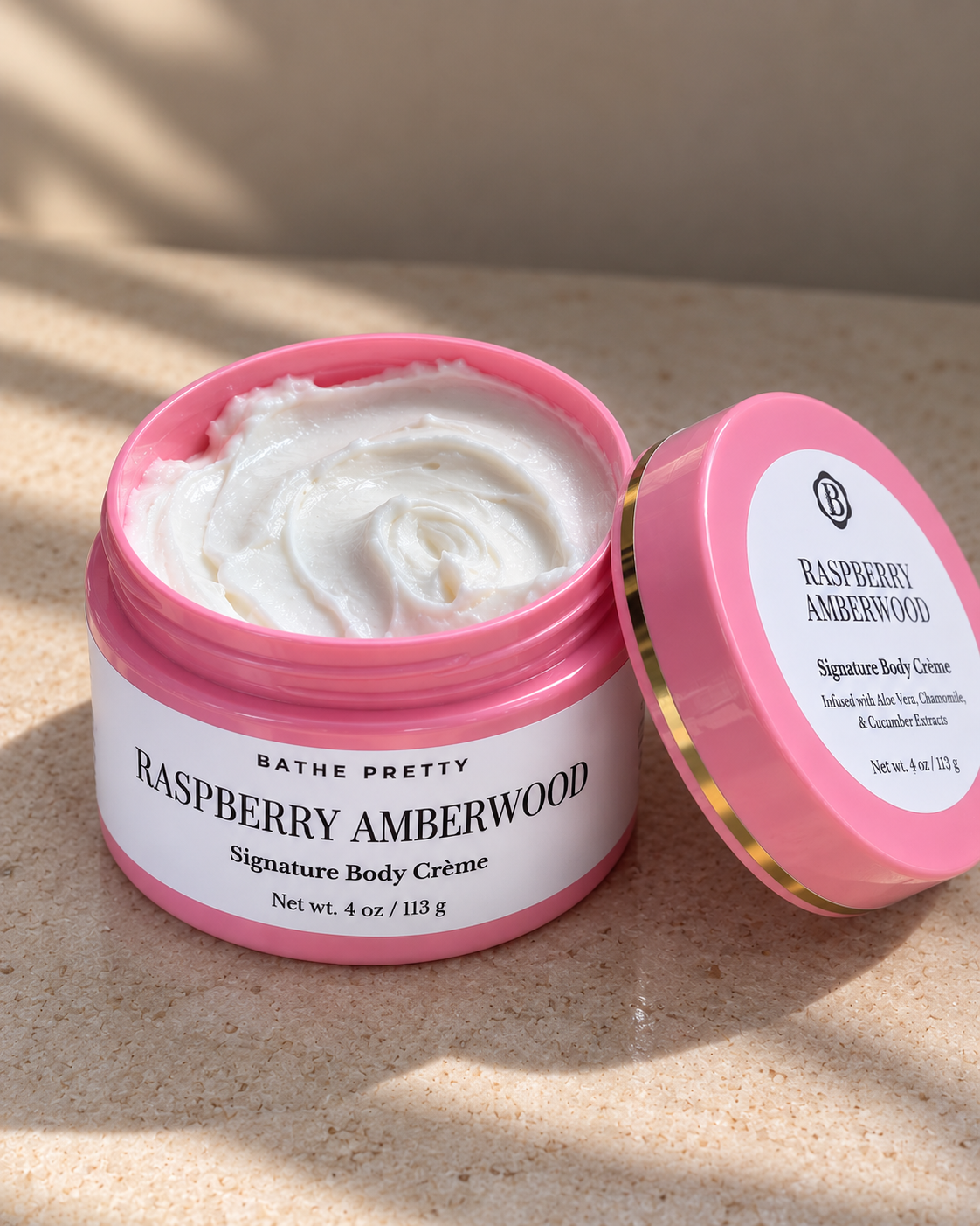 A jar of Bathe Pretty Raspberry Amberwood Signature Body Crème with its lid resting beside it on a textured beige surface, lit by warm natural sunlight.