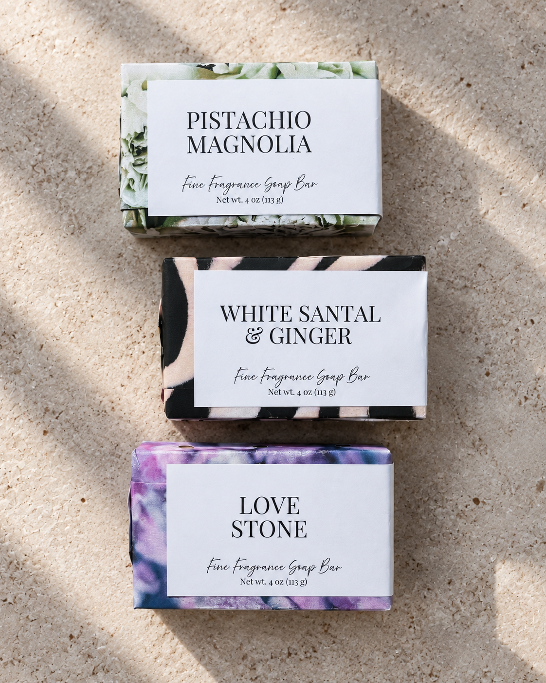 Three wrapped fragrance soap bars labeled Pistachio Magnolia, White Santal & Ginger, and Love Stone, arranged vertically on a textured beige stone surface with natural shadows.