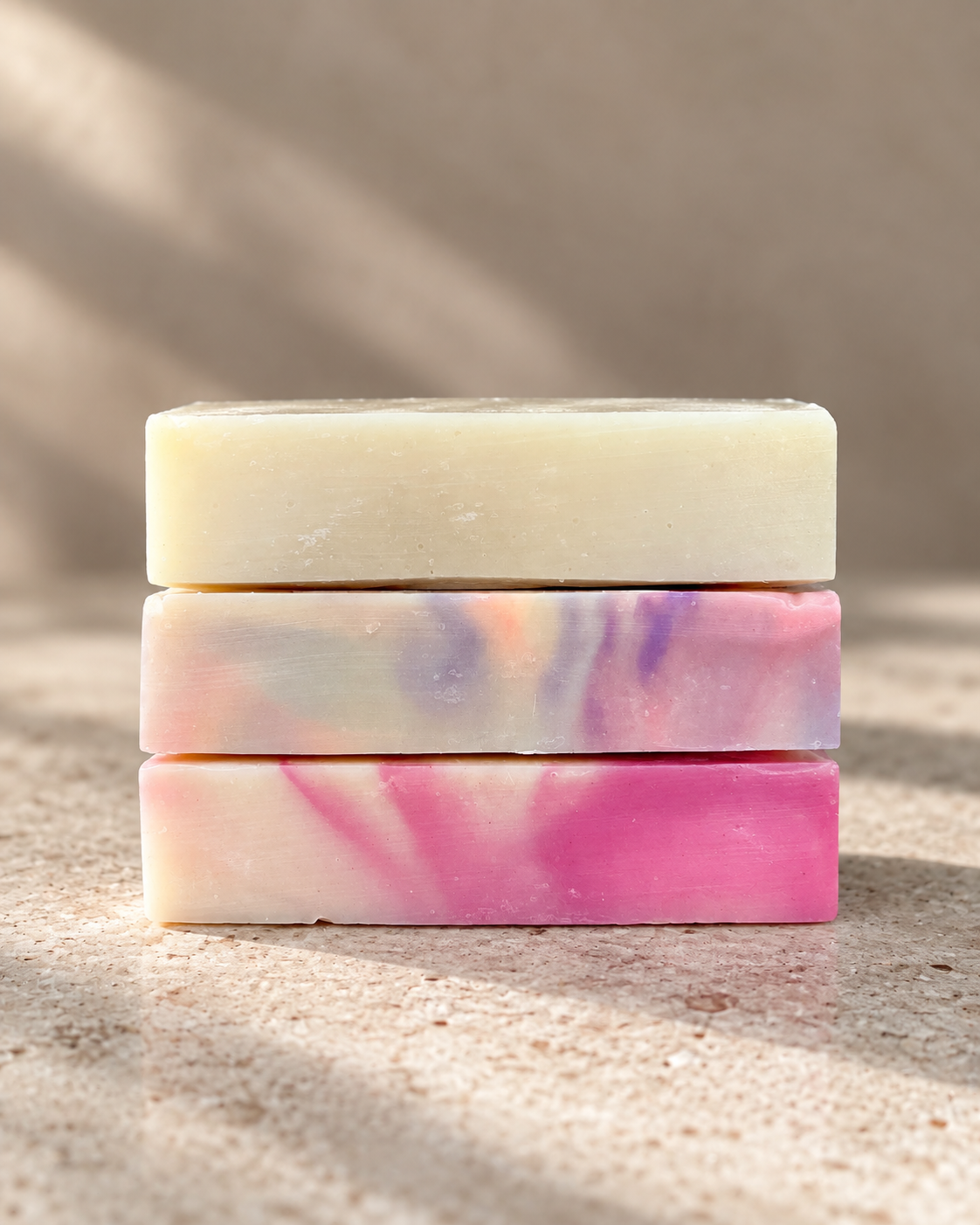 Three stacked rectangular bars of handmade soap on a sandy surface with soft shadows. The top bar is solid cream, while the middle and bottom bars feature a marbled swirl pattern of pink, purple, and cream.