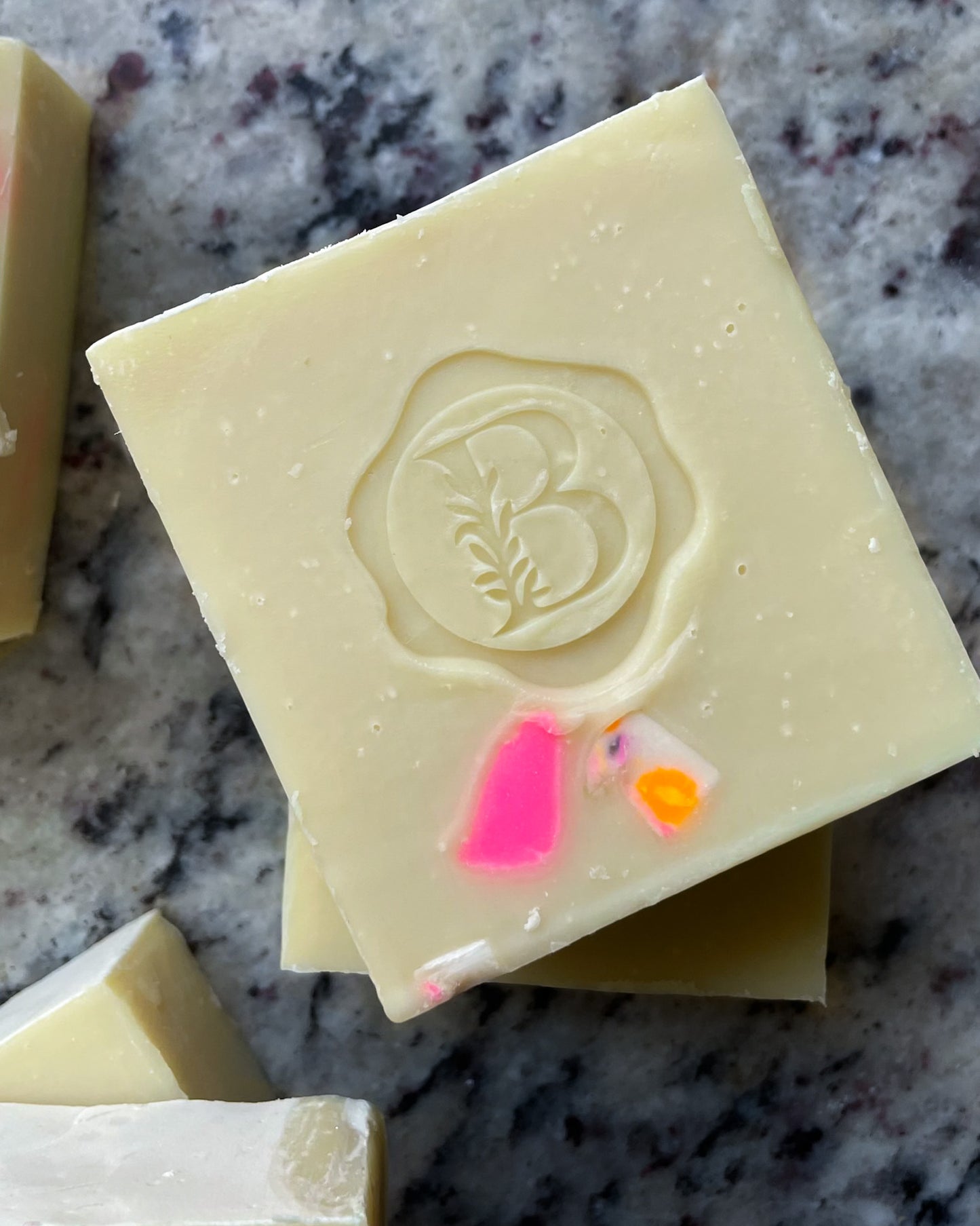 A close-up of a square bar of pale cream soap with a raised monogram 'B' and small bright pink and orange inclusions, resting on a gray marbled surface.