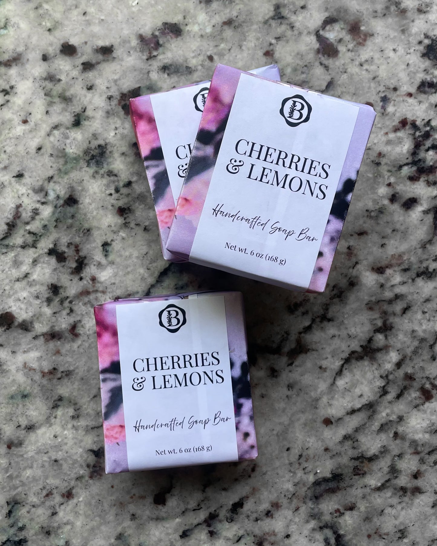 Three wrapped soap bars labeled 'Cherries & Lemons' with a white front label and pink floral sides, arranged on a speckled stone surface.
