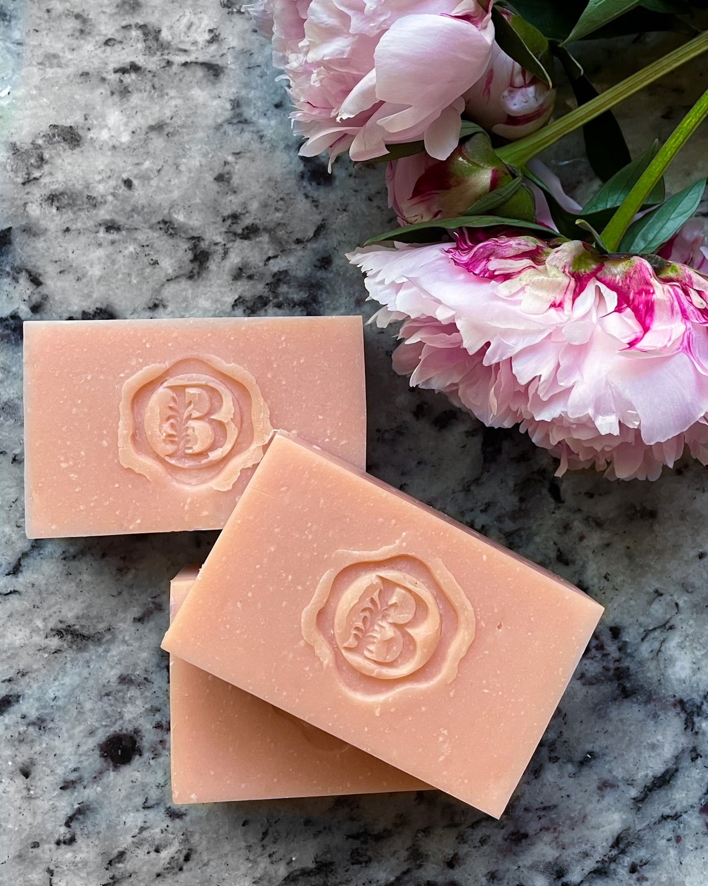 Three rectangular peach-colored soap bars stacked diagonally on a gray marbled surface, with pink peony flowers in the upper right corner. Each soap bar has an embossed circular monogram logo featuring a stylized letter 'B'.