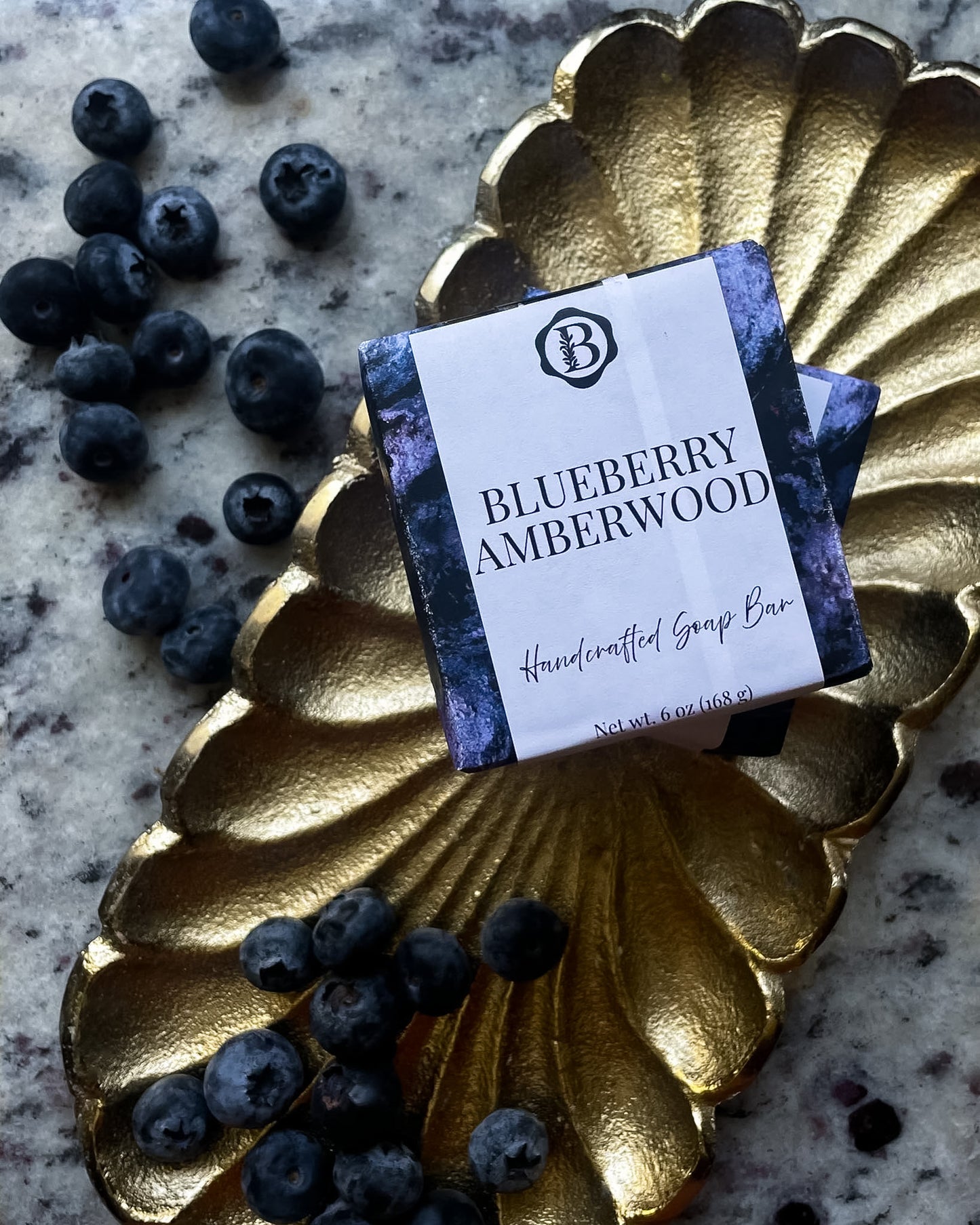 A packaged soap bar labeled 'Blueberry Amberwood' rests on a metallic gold scalloped dish with scattered blueberries on a speckled stone surface.