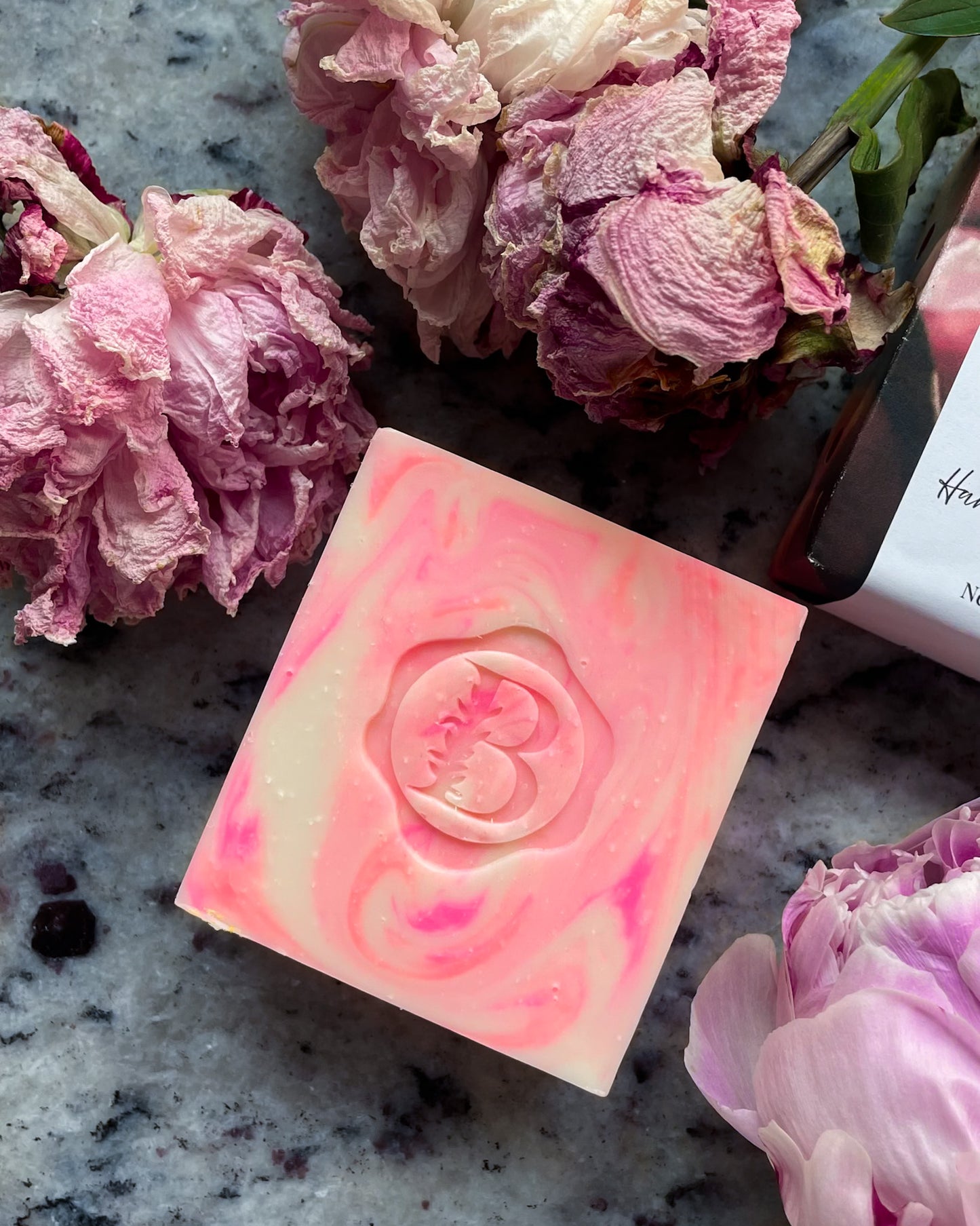 A rectangular bar of pink and cream marbled soap with a rose embossed in the center, styled on a gray marble surface with dried pink flowers and a bottle label nearby.