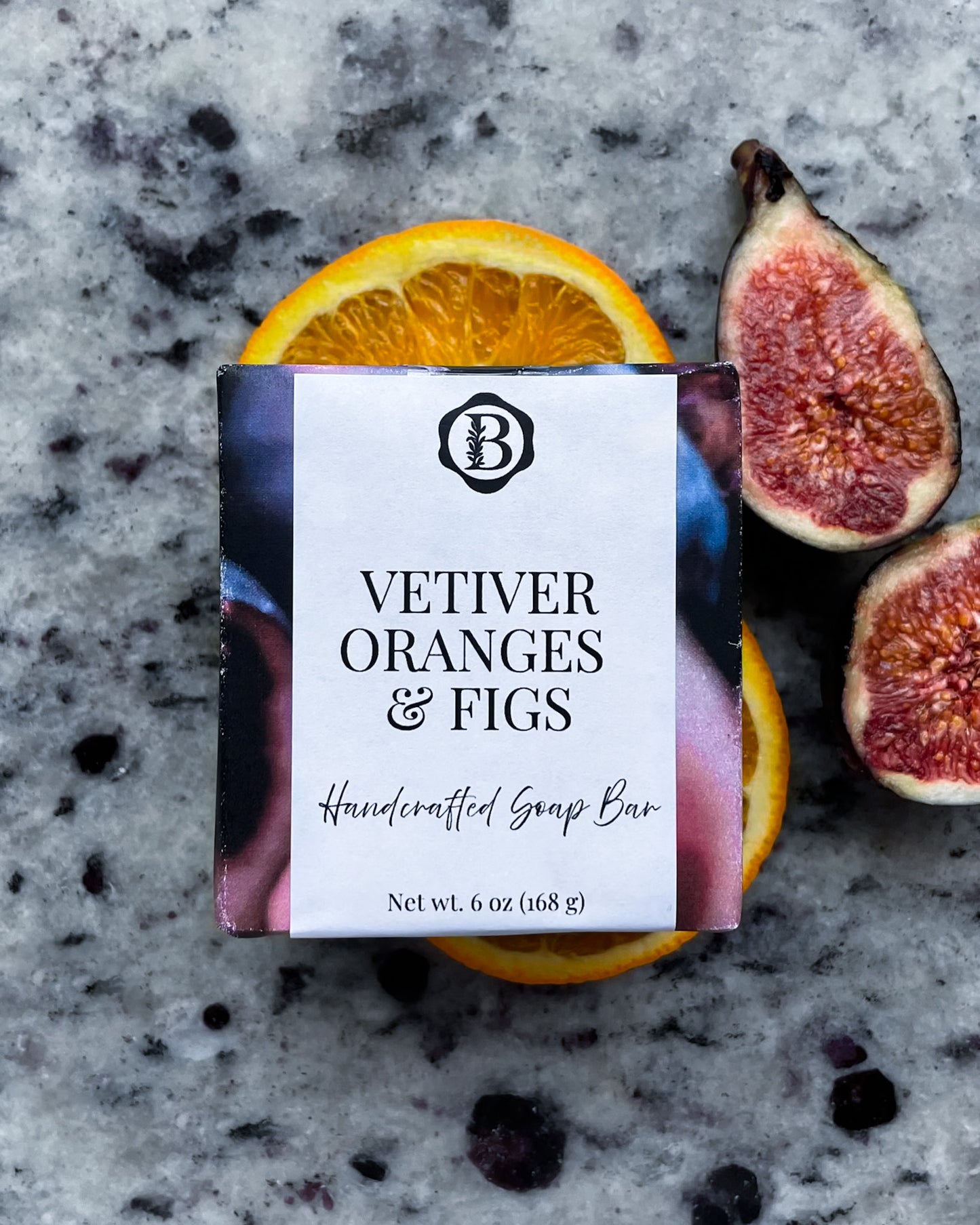 A wrapped  handcrafted soap bar labeled 'VETIVER ORANGES & FIGS' sits on a gray stone surface with sliced orange and fig halves around it. The wrapper has a white front label and colorful side panels, and no person or real environment is present beyond a styled flat-lay setup.
