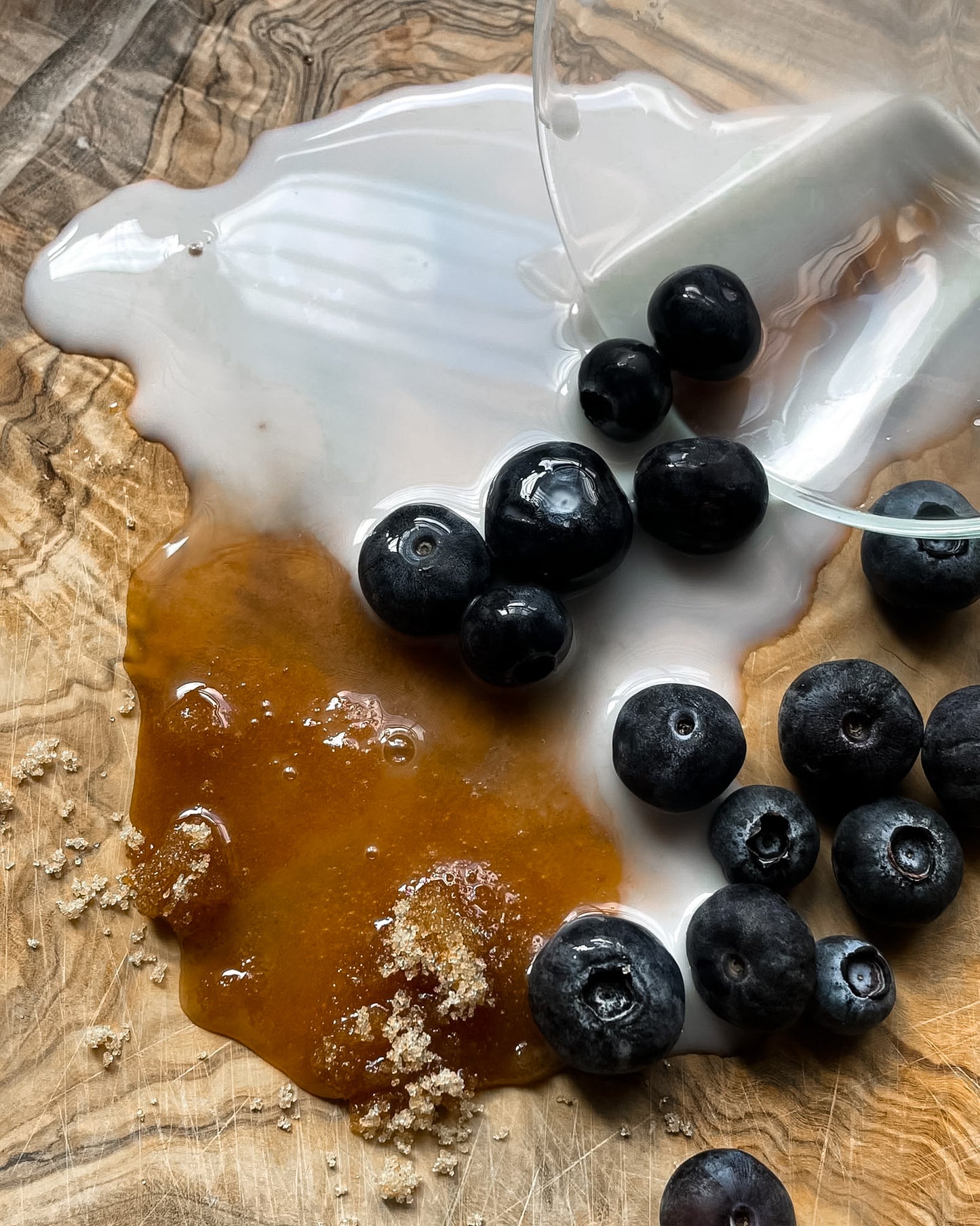 Blueberries and a glossy amber syrup pool on a wooden surface, with a white creamy liquid being poured over them from a clear bowl.