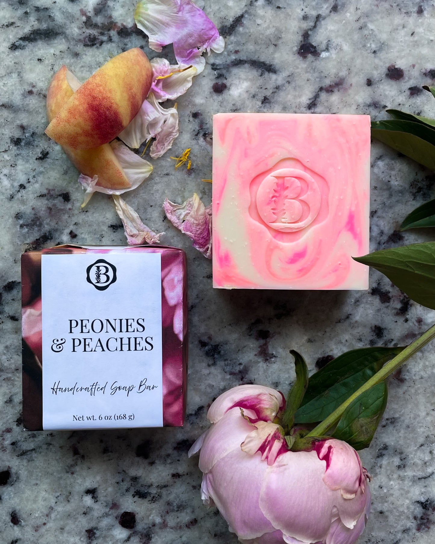 A handcrafted soap bar set displayed on a gray speckled stone surface with peony flowers. The set includes a pink and cream swirled soap bar with an embossed 'B' logo, its pink-and-white labeled wrapper, and loose peony petals and stems.