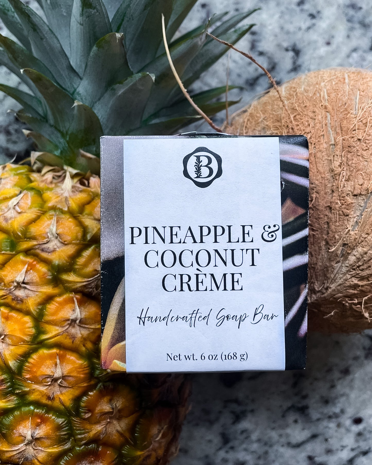 A wrapped handcrafted soap bar labeled 'PINEAPPLE & COCONUT CRÈME' placed on top of a pineapple and a coconut on a speckled gray countertop.