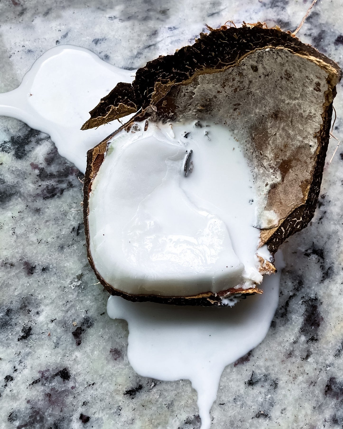A close-up of a broken coconut shell with white coconut cream or coconut oil pooled around it on a speckled gray stone surface.