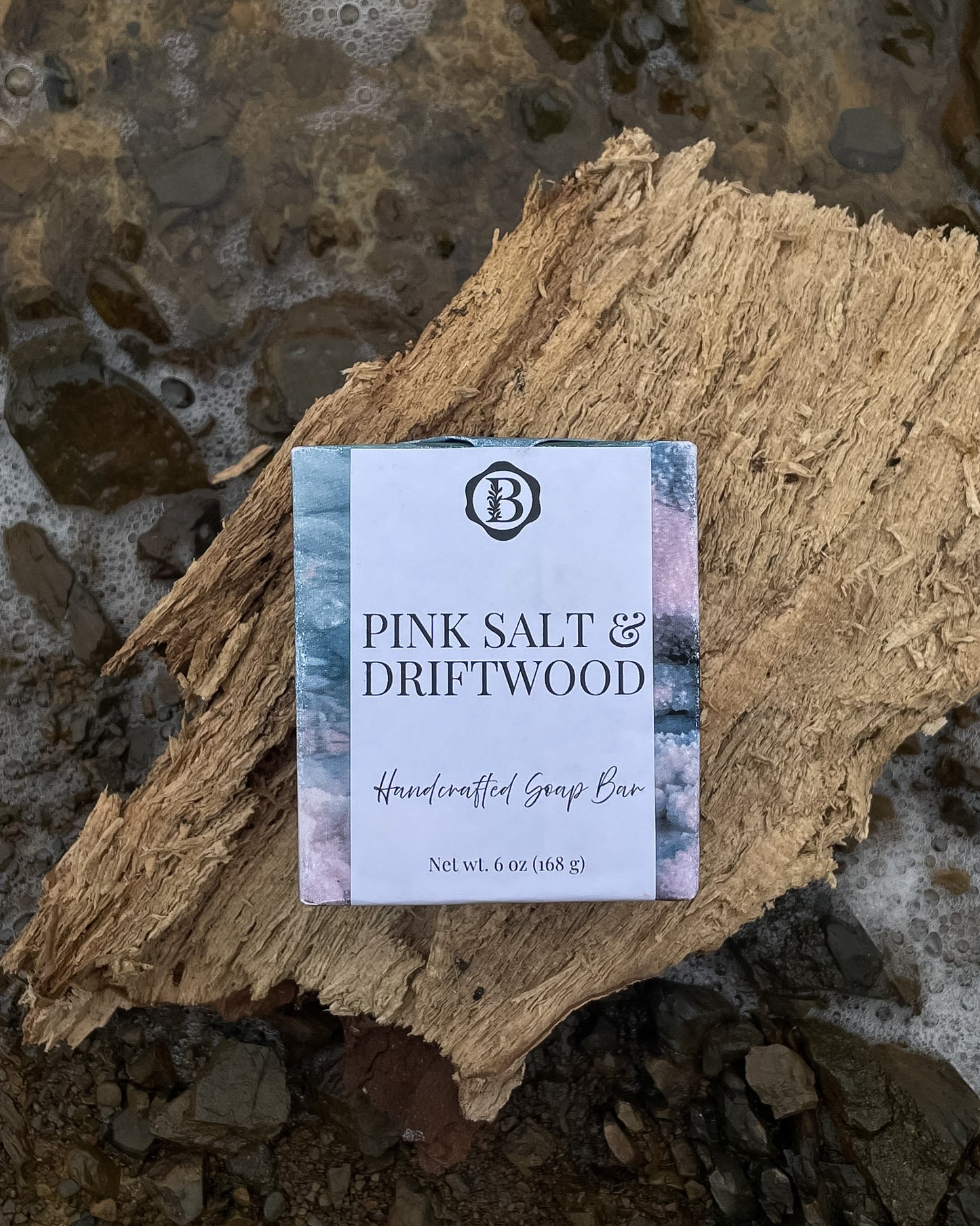 A rectangular bar of soap labeled 'PINK SALT & DRIFTWOOD' and 'Handcrafted Soap Bar' rests on a piece of rough driftwood over a rocky shoreline with water and foam visible around it. The soap wrapper has a white center with blue and pink watercolor-like side panels and a black "b' monogram logo at the top.