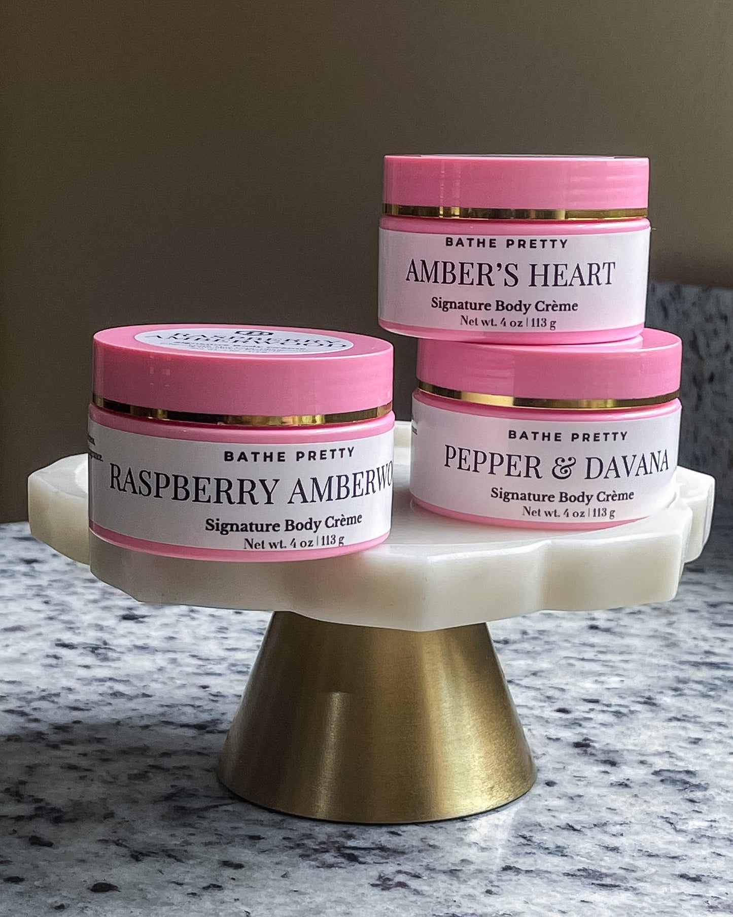 Three jars of Bathe Pretty Signature Body Creme displayed on a white and gold pedestal stand. The products include Raspberry Amberwood, Pepper & Davana, and Amber's Heart.