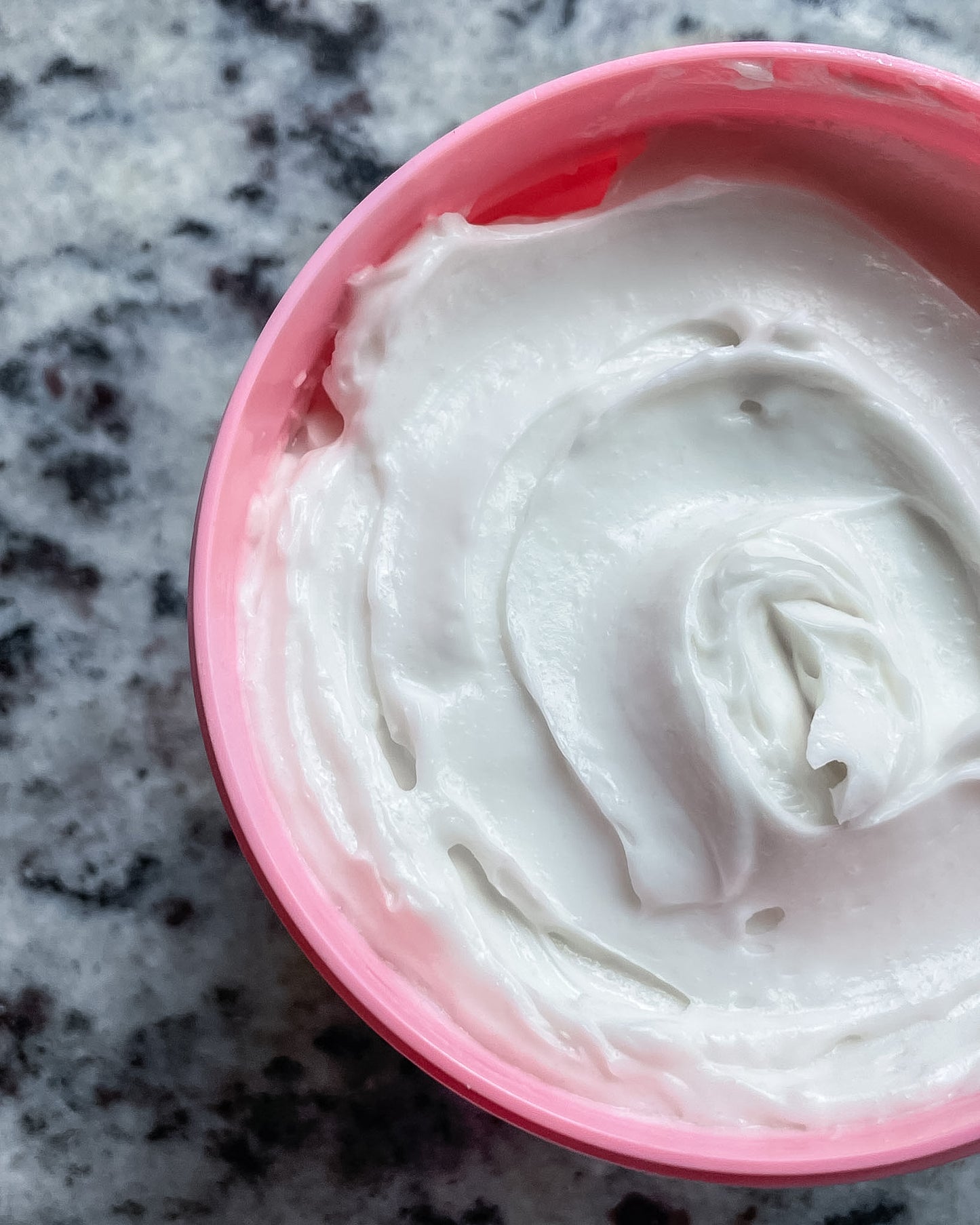 A close-up top-down view of a pink tub filled with thick white skin cream, set on a speckled gray countertop.