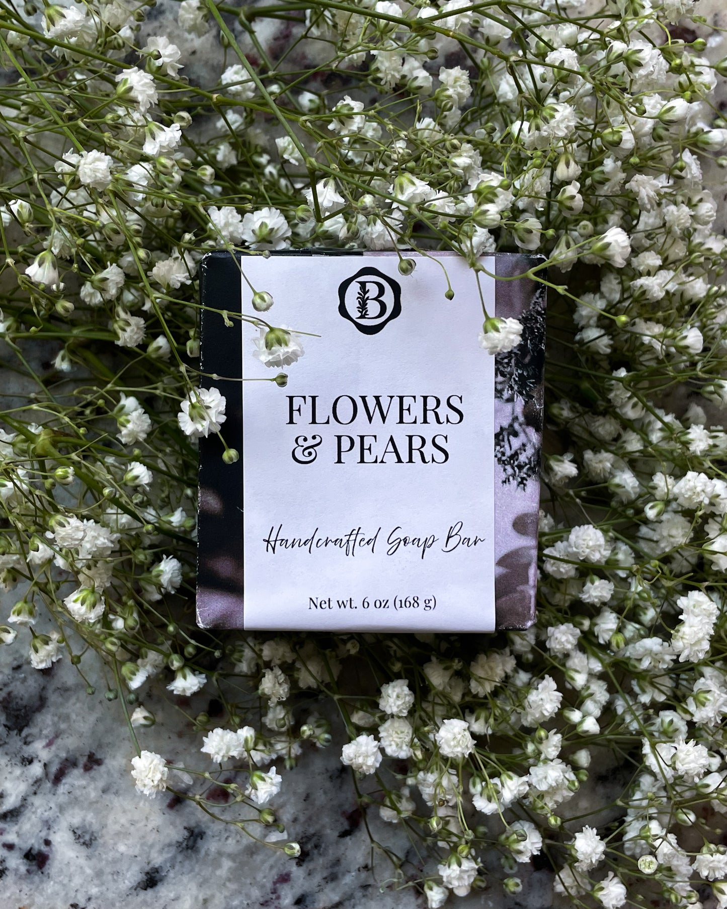 A handcrafted soap bar wrapped in a white paper label reading 'FLOWERS & PEARS' with a "b" monogram logo, placed on a bed of small white flowers over a gray stone surface.