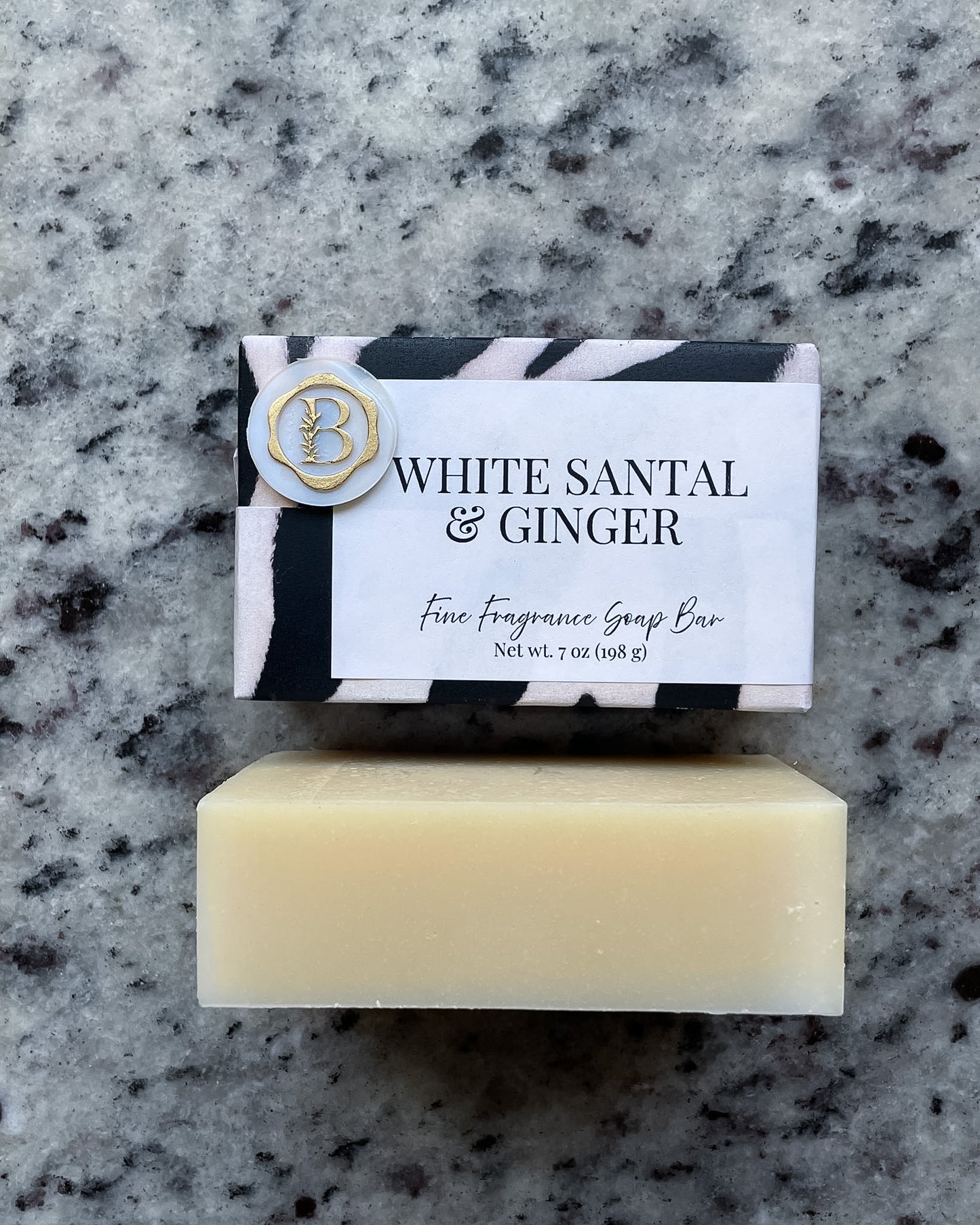 A bar of White Santal & Ginger fine fragrance soap bar placed on a gray speckled granite surface, with its black, white, and gold patterned paper packaged soap bar resting above it.