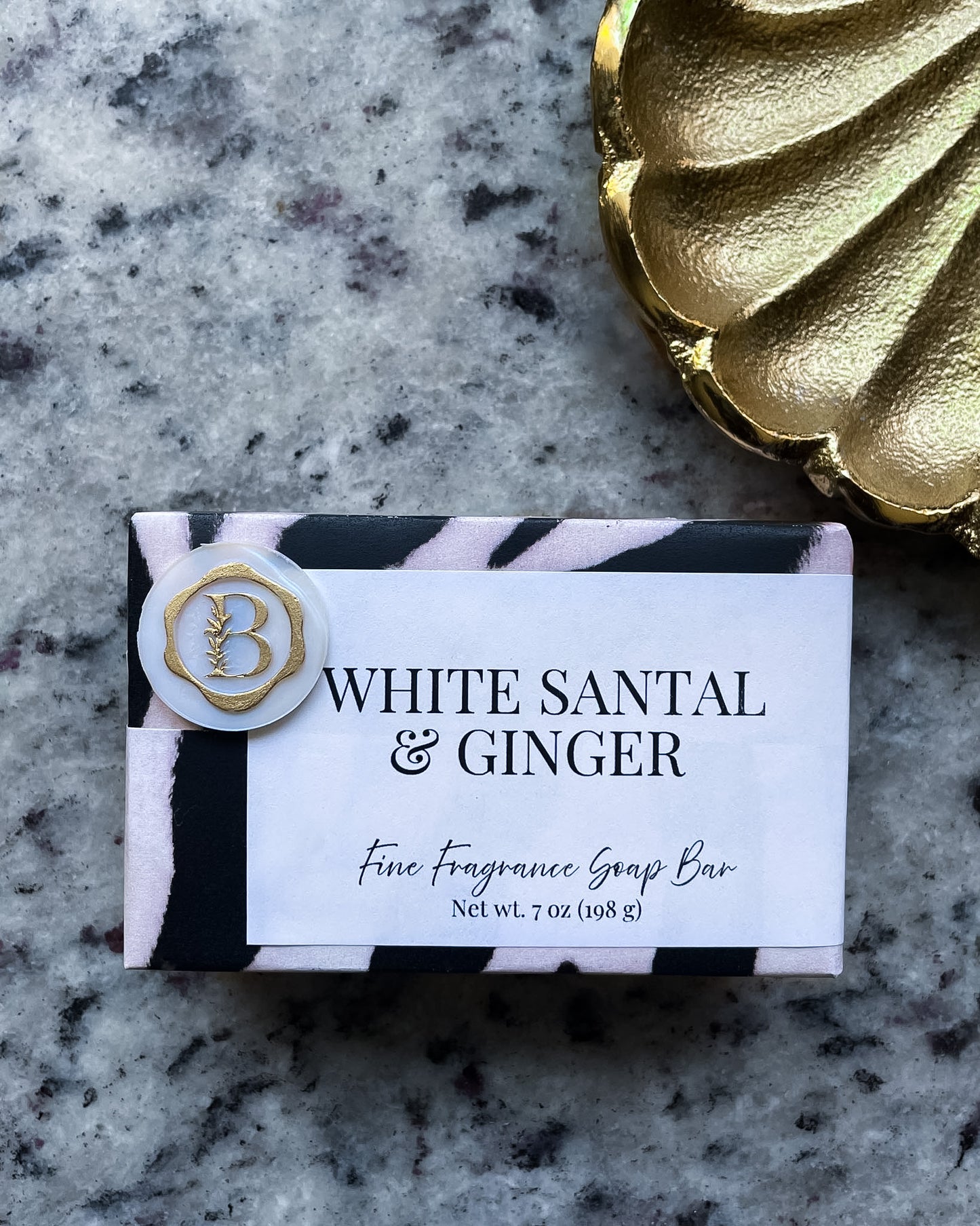 A wrapped fragrance soap bar labeled 'WHITE SANTAL & GINGER' with a white label, black and pale pink zebra-style border, and a round white seal with a gold monogram, placed on a gray marbled surface next to a gold decorative object.
