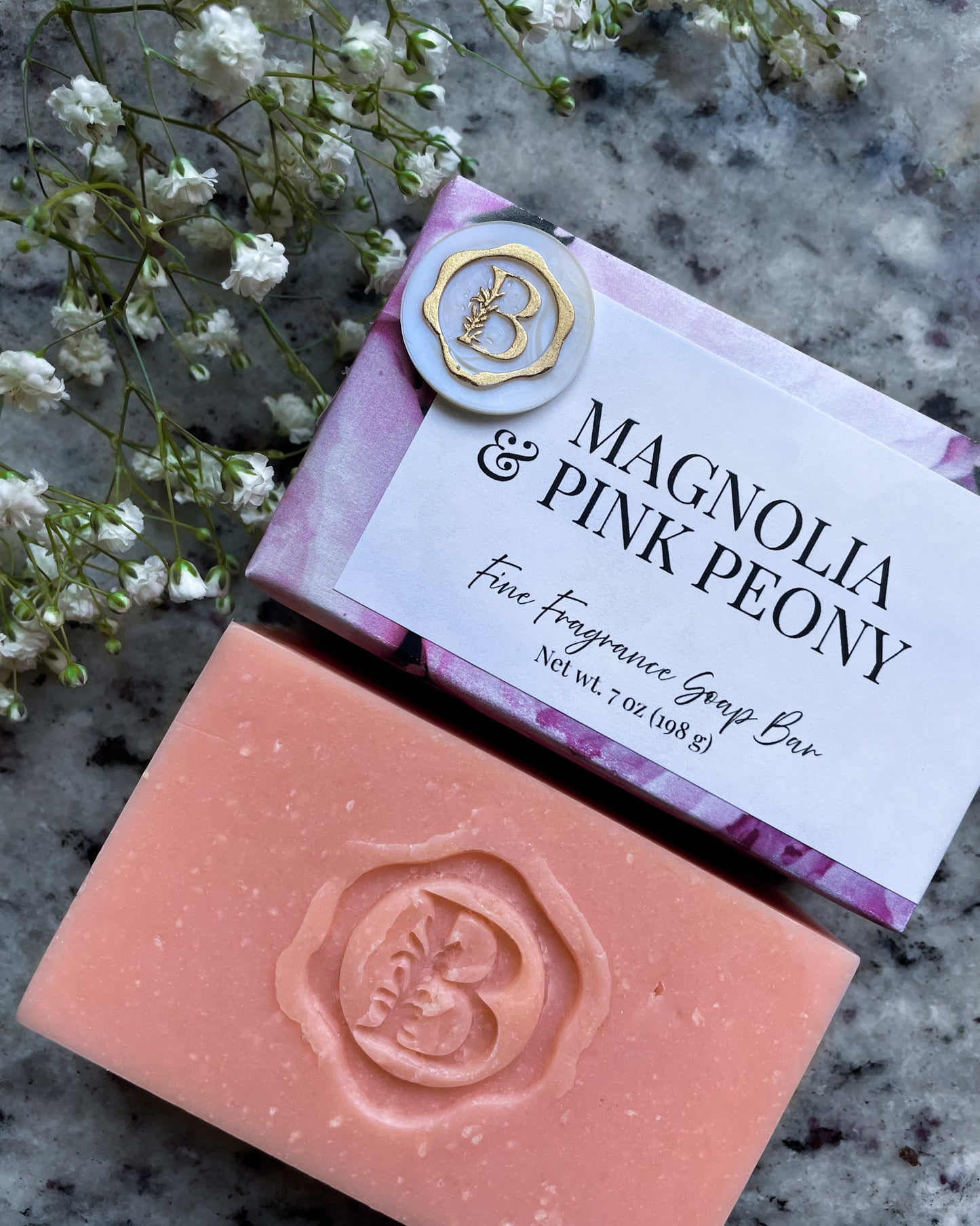 A bar of fine fragrance soap labeled 'Magnolia & Pink Peony' shown with its wrapped packaging on a stone surface, styled with small white flowers. The soap bar is peach-pink with an embossed logo, and the wrapper is lavender with a gold "b" seal.