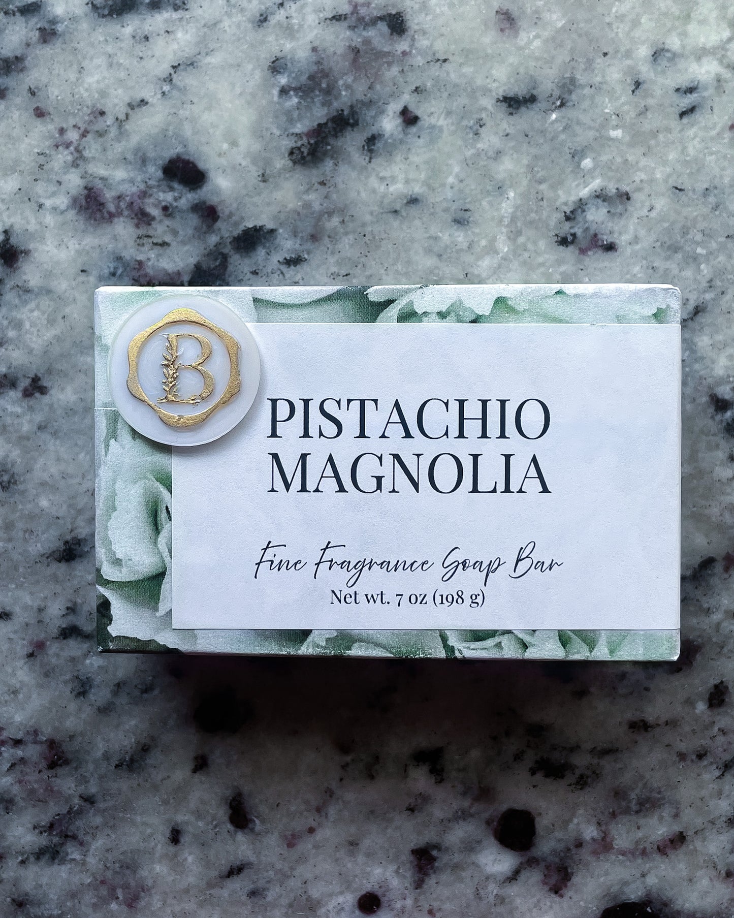 A wrapped  fragrance soap bar labeled 'Pistachio Magnolia' with a white front panel, green floral border, and a round gold-and-white "b" monogram seal, placed on a speckled gray stone surface.