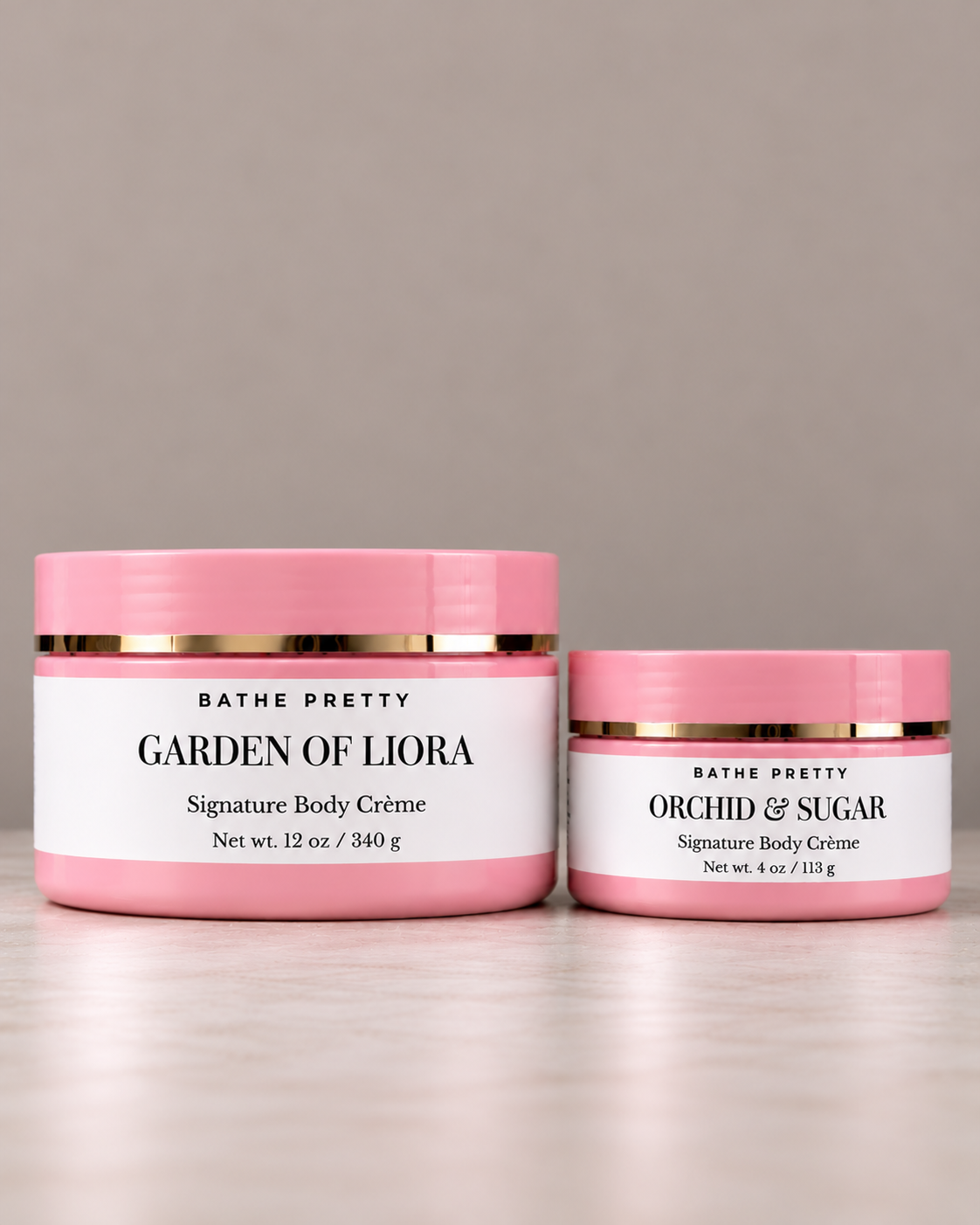 Two closed pink jars of Bathe Pretty Garden of Liora Signature body Crème in 12 ounces, next to a four ounce jar of orchid & sugar signature body crème  on a beige textured surface.